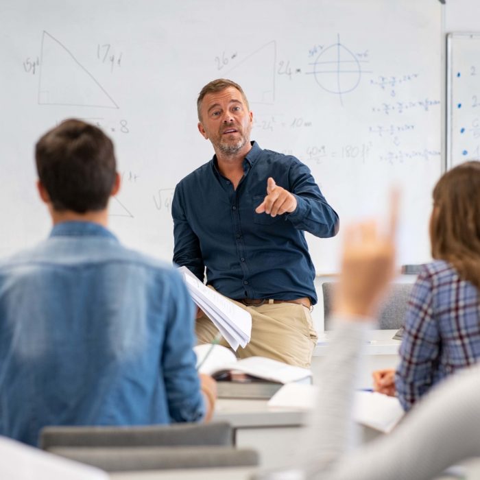 Professor pointing at college student with hand raised in classroom. Student raising a hand with a question for the teacher. Lecturer teaching in class while girl have a question to do during a math lesson.